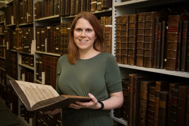 A woman with brown hair and a green dress stands in amongst medieval books.